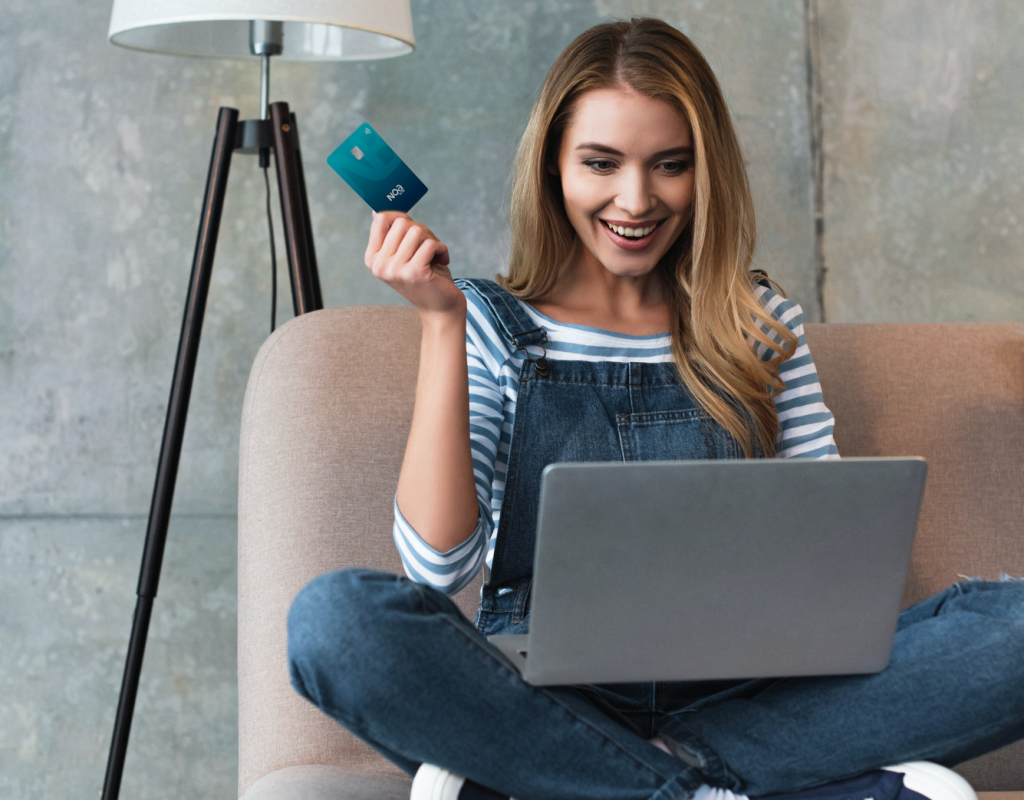happy-beautiful-woman-holding-credit-card-in-hand-and-sitting-on-sofa-with-laptop-noa-bank